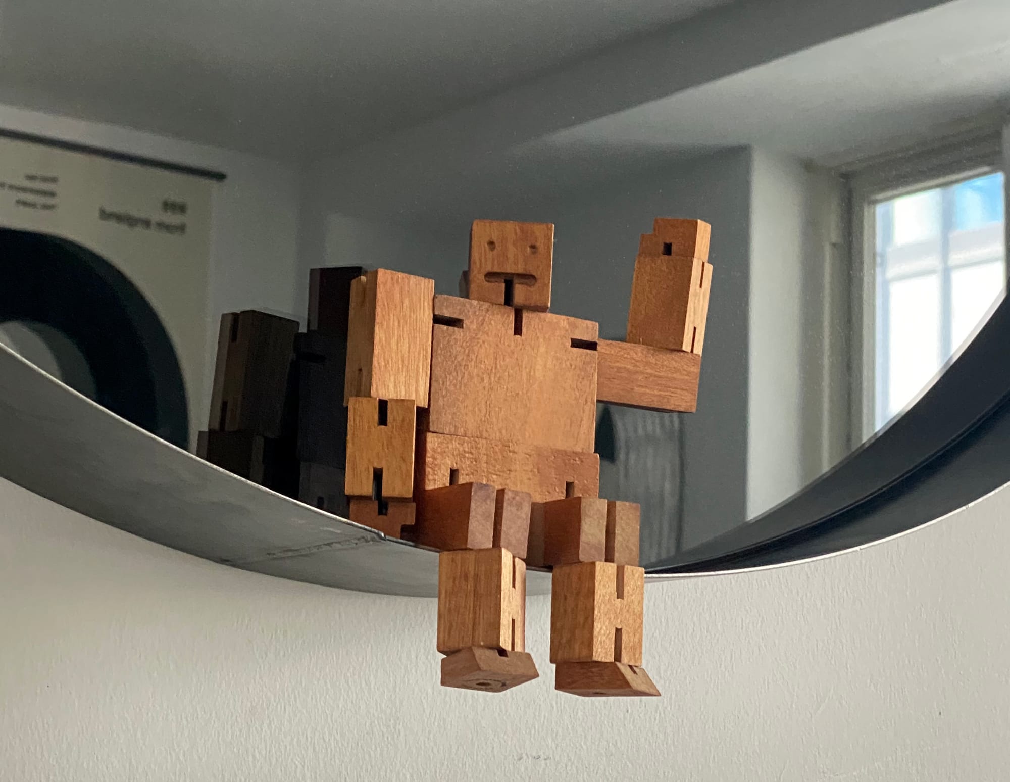 Wooden robot sitting in the lip of a circular mirror.