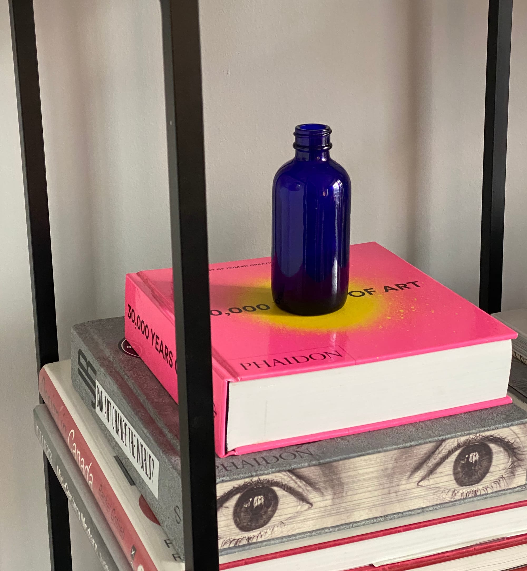Small blue bottle on a hot pink book that has a yellow spraypaint-like marking in the centre of its front cover.