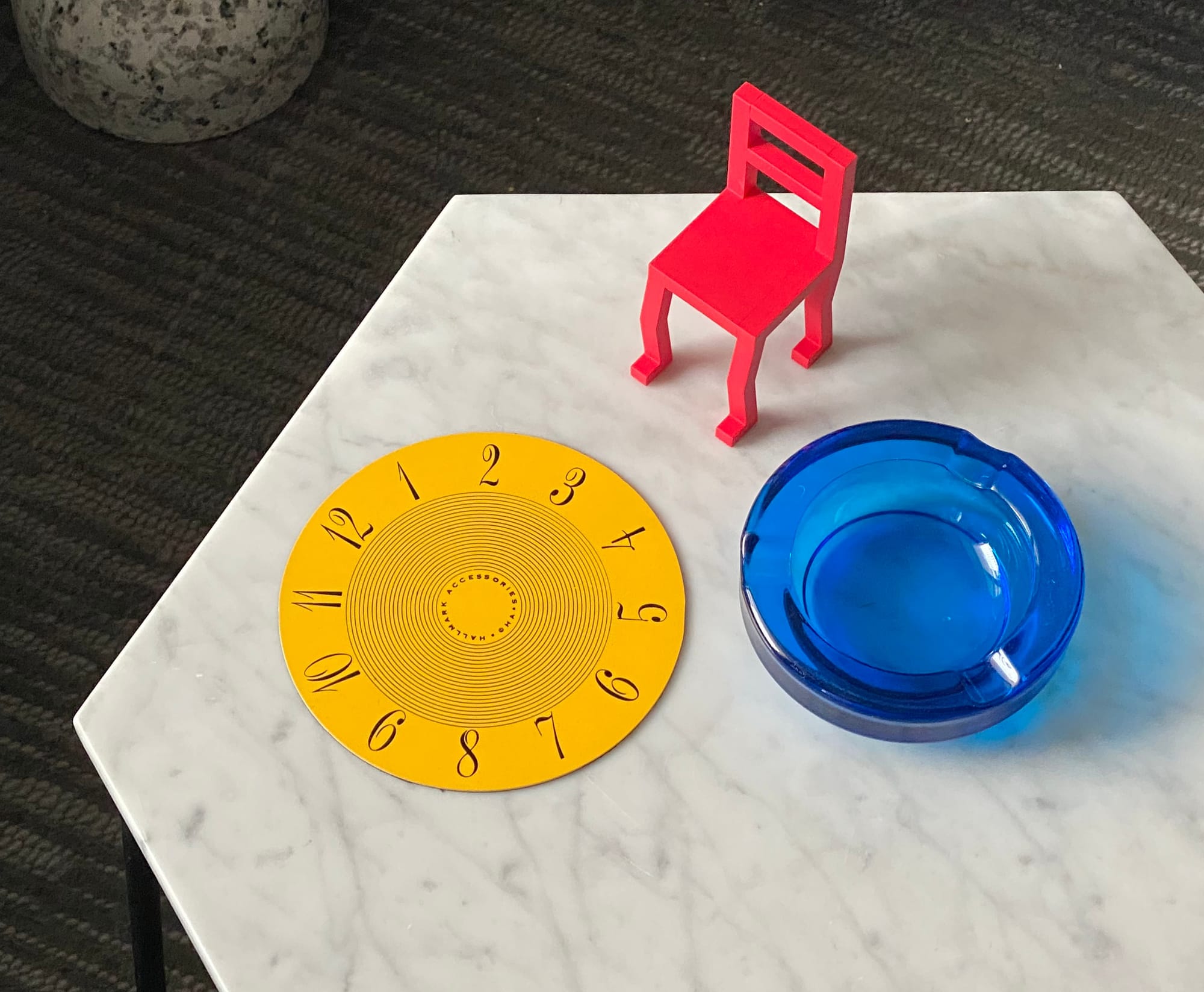 Yellow face from a clock, together with blue ashtray and miniature red chair, all on a marble tabletop.