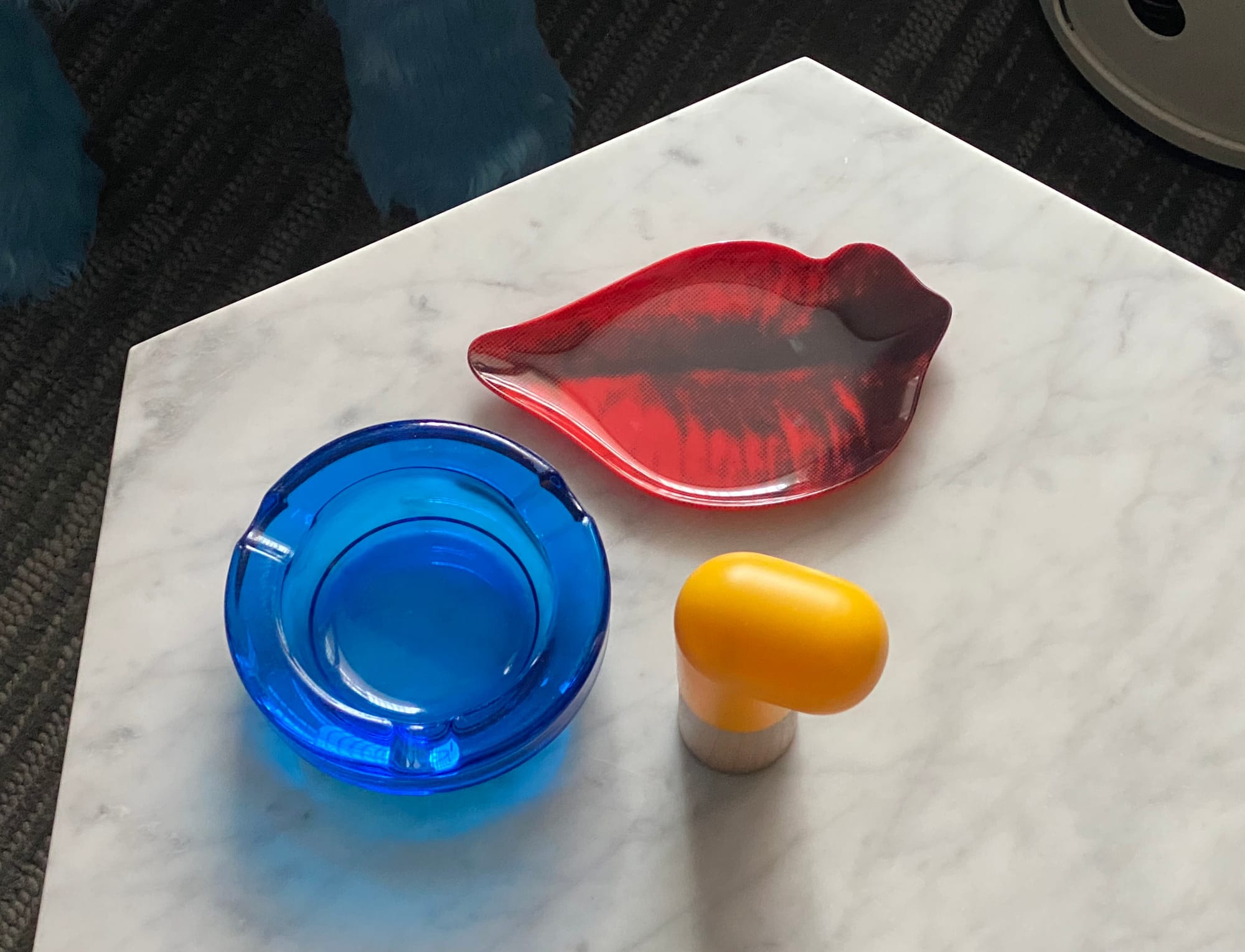 Tray in the shape of red lips, together with a blue ashtray and yellow plastic item resting on a wooden block, all on a marble tabletop.