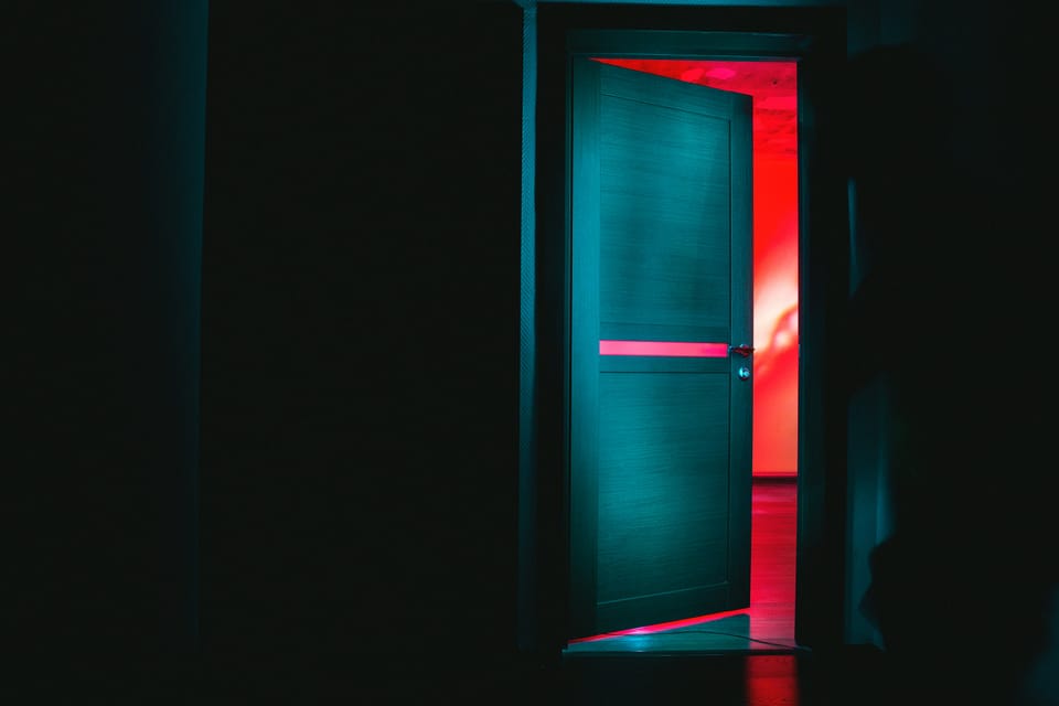 Dark room with an open door revealing a red room beyond the threshold.